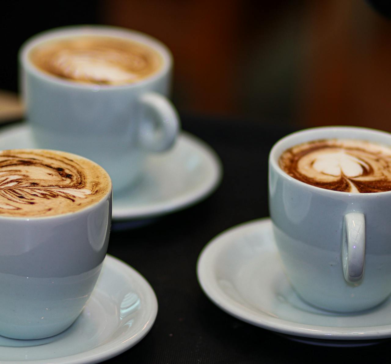 heros-img Three cappuccinos with intricate latte art in white cups on saucers, showcasing coffee artistry.